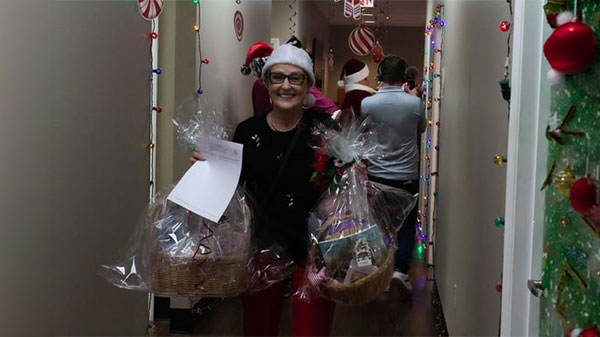 A festive woman in a Santa hat carries two decorated gift baskets down a holiday-themed hallway with cheerful decorations, celebrating the season.