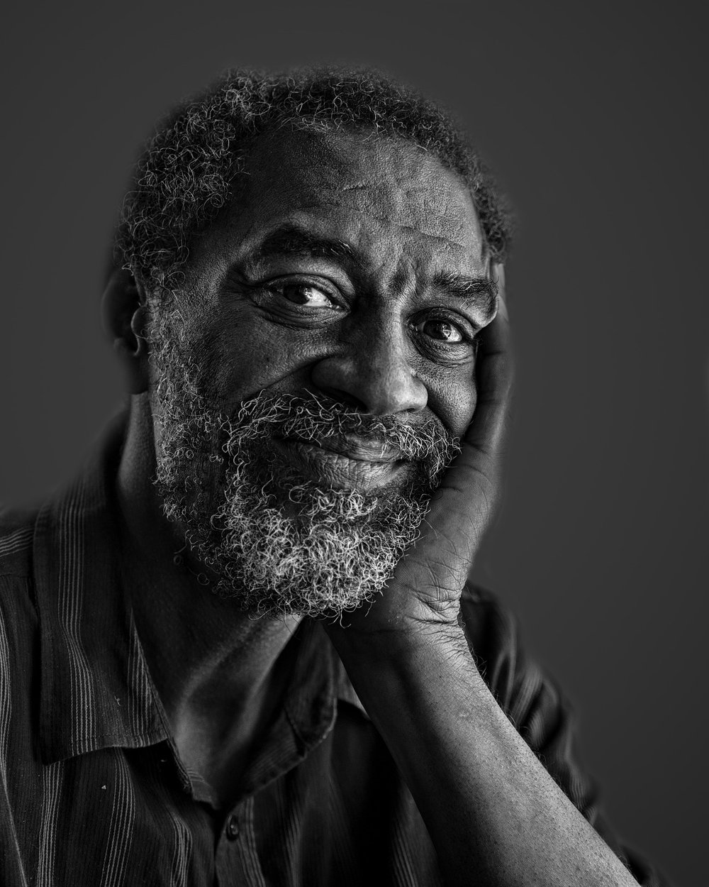 A smiling older man with a beard rests his chin on his hand, exuding warmth and wisdom against a dark background.