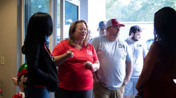 A group of four people converse indoors, with one woman in a red shirt gesturing as she speaks. The scene captures a friendly discussion among attendees.